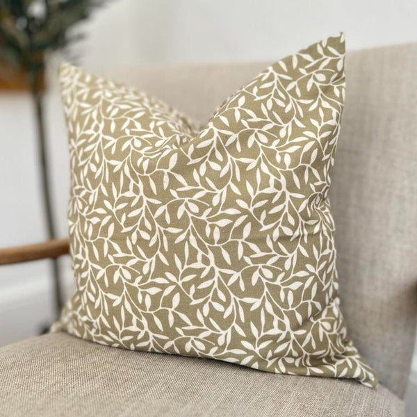 square cotton cushion in olive green with an all over cream coloured olive leaf print sat on a cream armchair.