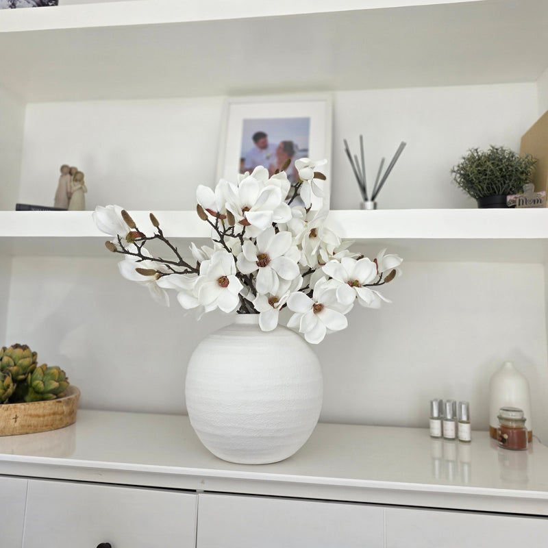 Faux White Magnolia Stems sat inside a white round ceramic vase sat on a white sideboard in a living room.