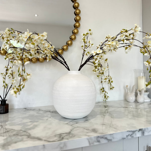 round white ceramic vase with a narrow short neck. White blossom stems pouring out of the neck. All sat on a kitchen island.