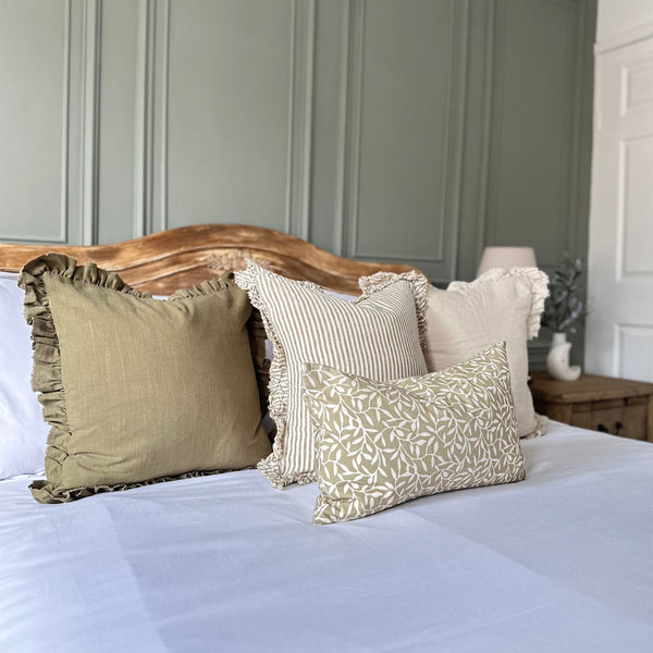 set of 4 cushions with olive tones, stripes and leaf rpints, sat on a bed with white bedding and a panelled green wall as background.