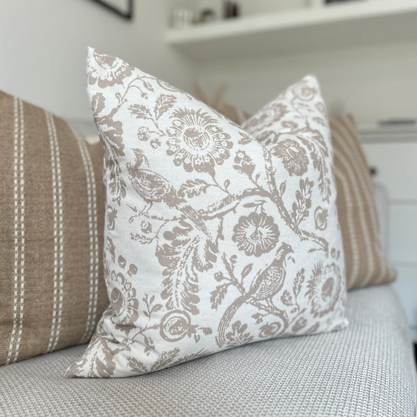 square cream cotton cushion with a large beige floral all over pattern with flowers, leaves and peacocks in beige. Sat on a cream sofa.