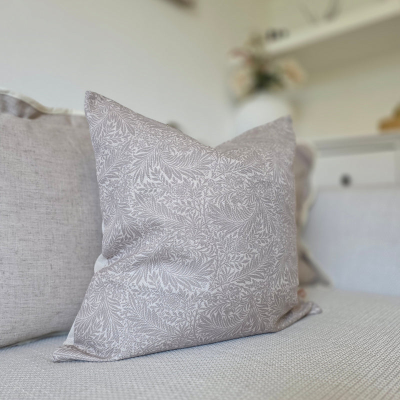 Clos e up of the Lillian greige leaf print cushion sat in front of our scalloped edge greige cushion on a cream sofa in a living room
