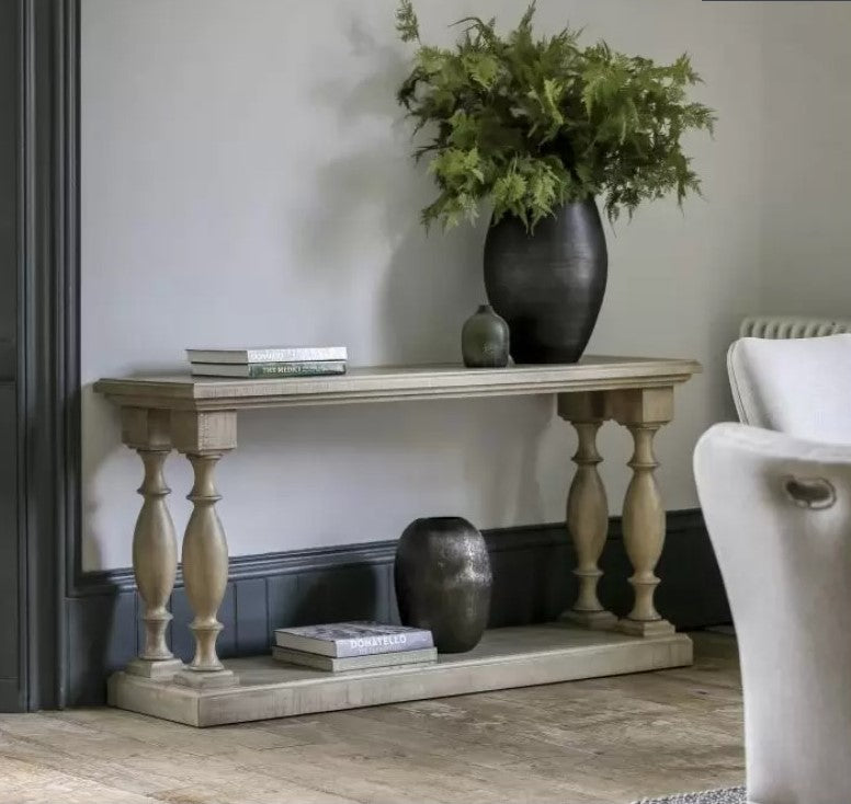 West Coast Accent Wooden Console Table with a large black vase with green foliage on top and books with a small vase sat on the bottom wooden base of the console table.