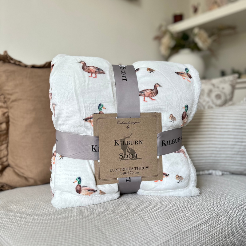 Watercolour Sherpa Duck Print Blanket. White soft and fluffy background with all over duck and duckling watercolour print. sat on a cream sofa