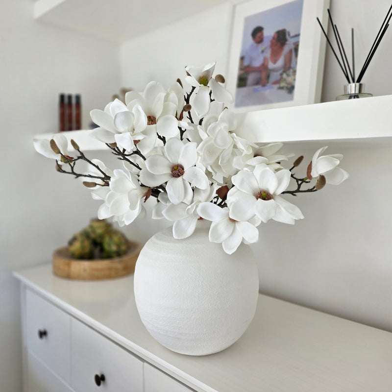 3 magnolia faux stems sitting in a white round vase on a white sideboard in a living room