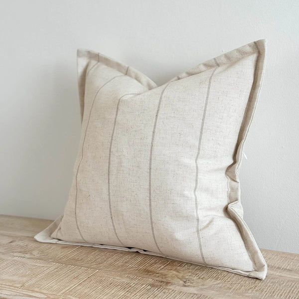 Flanged edge square cotton cream cushion with a slim all over vertical beige stripe. Sat on a wooden side with a plain white wall background for contrast