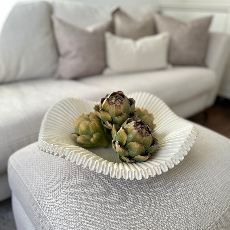 White scalloped edge decorative large bowl, with decorative green artichokes inside. Sat in the centre of a white sideboard