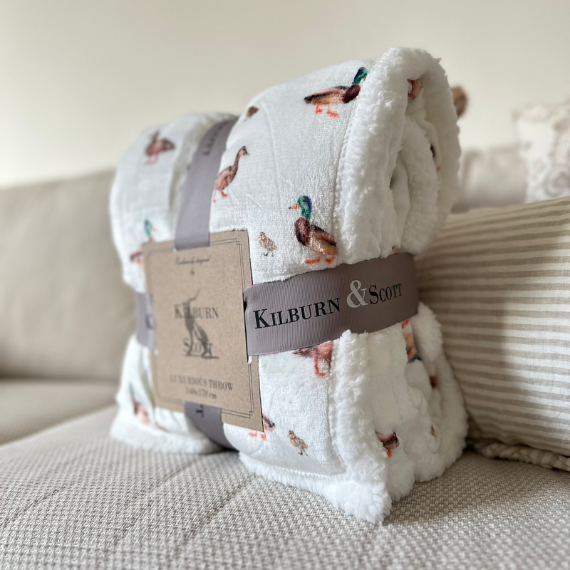 Side view of the watercolour. Sherpa Duck Print Blanket. White soft and fluffy background with all over duck and duckling watercolour print. sat on a cream sofa