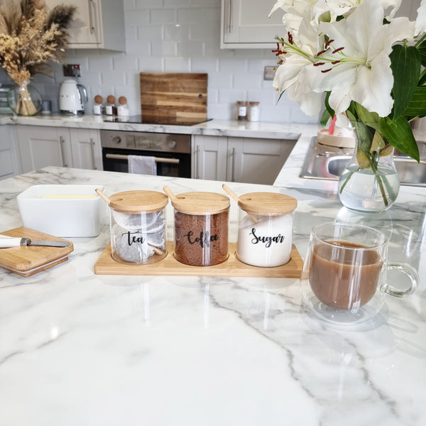 Set of 3 Glass jars with bamboo spoons and lid and customisable black wording for storing tea coffee and sugar sat on wooden bamboo tray